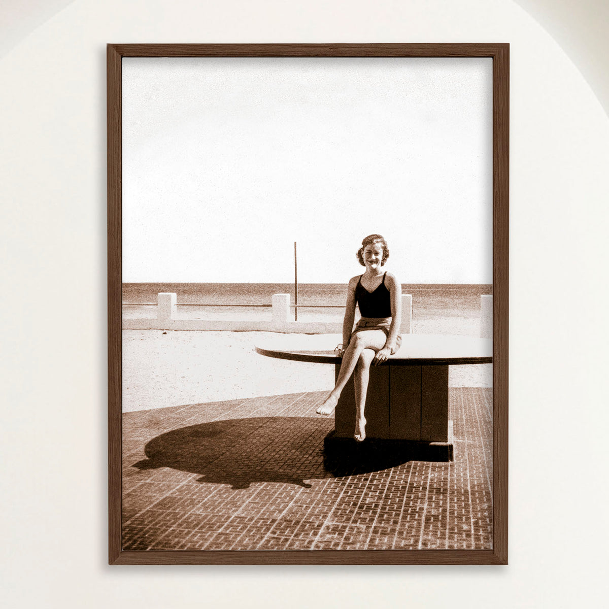 Sepia photograph of a 1950s woman seated by the sea at Playa Cojimar in vertical format, framed in oak. Close view.