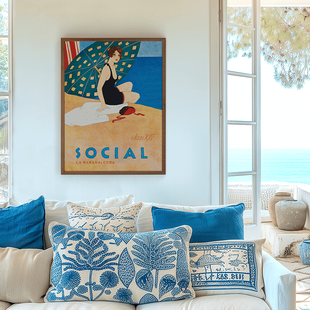 Framed 1920s Art Deco illustration with visible artist details of a woman in a black bathing suit on a Havana beach under a green umbrella, displayed above a white sofa in a bright coastal living room with French doors and a sea view.