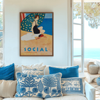 Framed 1920s Art Deco illustration with visible artist details of a woman in a black bathing suit on a Havana beach under a green umbrella, displayed above a white sofa in a bright coastal living room with French doors and a sea view.